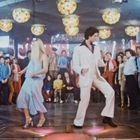 Eine Menschenmenge in einem Club, ein Paar tanzt in der Mitte; bunte Lichter an der Decke.