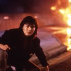 Eine Person kniet auf einer Straße, im Hintergrund brennt etwas intensiv.