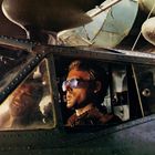 Zwei Männer im Cockpit eines alten Flugzeugs; einer trägt eine Sonnenbrille. Sichtbare Nieten und Technik im Hintergrund.