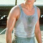 Ein junger Mann in einem verschwitzten grauen Tanktop steht fokussiert auf einem Sportplatz. Er trägt graue Shorts.