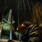 Keine erkennbaren Texte. Eine bedrohliche, humanoide Kreatur mit einem pyramidalen Helm steht hinter Gittern, während eine Person aufblickt.