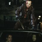 Ich kann keinen spezifischen Text identifizieren. Eine Person mit langen Haaren und blutendem Gesicht steht bedrohlich auf einem Auto. Zwei Menschen sitzen im Auto: Der Fahrer wirkt konzentriert, der Beifahrer hat blutende Verletzungen im Gesicht. Es ist Nacht.