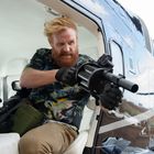 Ein Mann mit rotem Bart in einem Hubschrauber hält eine große Waffe und trägt ein bunt gemustertes Hemd.