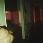 Ich kann keine Texte erkennen. Ein Mann steht gebeugt vor einer Wand, in einem düsteren Flur mit rotem Licht im Hintergrund.