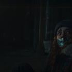 Keine sichtbaren Texte. Eine Person mit gefesseltem Mund sitzt verängstigt in einem dunklen Raum.