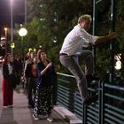Ein Mann klettert nachts ein Geländer, während eine Gruppe auf dem Bürgersteig schaut. Beleuchtete Straße im Hintergrund.