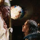 Ein Priester in prachtvollen Gewändern steht vor einer Person mit langer, nasser Frisur in einer kunstvollen Umgebung.