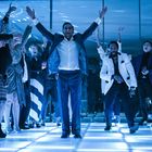 Eine Gruppe feiert auf einer beleuchteten Tanzfläche, zentral eine Person mit erhobenen Armen. Elegante Kleidung, freudige Gesichter.