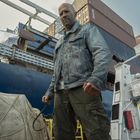 Eine Person in Jeansjacke vor Containern auf einem Frachter, hält einen gebogenen Metallstab.