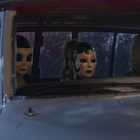 Drei verhüllte Personen in einem Fahrzeug, alle tragen unheimliche Masken, nachts auf der Rückbank sitzend.