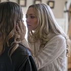 Zwei Frauen stehen sich nahe gegenüber, eine in einem weißen Pullover berührt das Gesicht der anderen Frau mit langen Haaren.