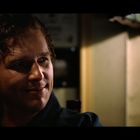 Ein Mann mit lockigem Haar lächelt leicht. Er ist in warmes, schummriges Licht getaucht, umgeben von dunklen, unscharfen Hintergründen.