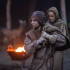 Keine Texte vorhanden. Eine junge Person in altertümlicher Kleidung steht im Fokus, umgeben von unscharfen Menschen und einem Feuer.