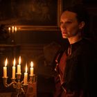 Kerzenständer mit brennenden Kerzen beleuchtet eine Frau in historischer Kleidung in einem gedämpften, eleganten Raum.