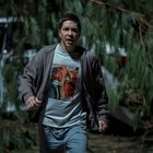 Ein Mann läuft durch eine bewaldete Gegend, trägt einen grauen Hoodie und ein Comic-T-Shirt mit mehreren Panels.