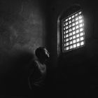 Person blickt aus einem Gitterfenster in einem düsteren Raum, Licht fällt durch das Gitter auf das Gesicht.