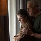 Ein älteres Paar steht nah beieinander und schaut ernst aus einem Fenster. Eine Lampe links beleuchtet den Raum.