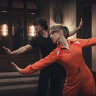 Zwei Tänzer in Bewegung, Frau in auffälligem roten Anzug, Mann in Schwarz. Elegante Armhaltungen, warme Umgebung.