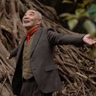 Ein lachender Mann in einem braunen Anzug mit rotem Schal breitet die Arme aus, im Hintergrund dicke Baumwurzeln.