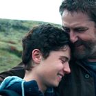 Ein Mann und ein Junge umarmen sich lächelnd auf einem grasbewachsenen Hügel.