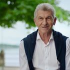 Es gibt keinen Text. Ein älterer Mann in weißem Hemd sitzt vor einem Baum. Im Hintergrund ist ein See zu sehen.