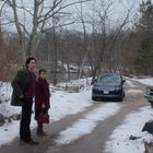 Zwei Personen in Winterkleidung stehen auf einem verschneiten Weg neben einem Auto, umgeben von kahlen Bäumen.