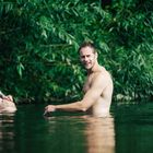 Im Wasser stehende Männer, einer mit Tätowierungen. Im Hintergrund grüne Blätter.