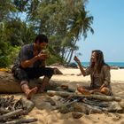 Ein Mann und eine Frau sitzen am Strand. Der Mann trinkt aus einer Kokosnuss, die Frau hält etwas Hoch. Im Hintergrund Meer.