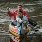 Zwei Personen paddeln fröhlich in einem bunt bemalten Kanu über einen ruhigen Fluss.