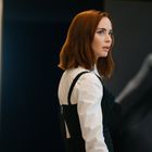 Eine Frau mit roten Haaren in weißer Bluse vor einem Schwarz-Weiß-Foto einer tanzenden Frau im Hintergrund.