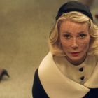 Keine erkennbaren Texte. Eine blonde Frau mit Schleier blickt nach oben, umgeben von unscharfen Personen.