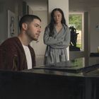 Ein Mann spielt Klavier, während eine Frau mit verschränkten Armen daneben steht. Im Hintergrund ist eine Statue sichtbar.