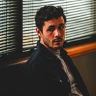 Ein Mann mit lockigem Haar sitzt nachdenklich vor Jalousien. Dezente Beleuchtung und entspannte Pose.