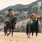 Zwei Personen auf Pferden tragen Cowboyhüte in einer bergigen Landschaft mit kahlen Bäumen und Gras.
