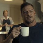 Ein Mann in einem dunkelblauen Shirt hält eine Tasse, während im Hintergrund zwei Personen in einer Küche stehen.