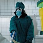 Auf einem Kanister steht "THE STUFF". Eine Person in OP-Kleidung und Maske hält einen Schlauch. Tiles im Raum.