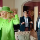 Eine Frau in grünem Outfit mit Hut hält ihn fest. Zwei Männer stehen im Hintergrund. Wohnzimmer mit heller Einrichtung.
