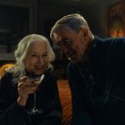 Eine ältere Frau mit grauem Haar hält ein Cocktailglas. Sie lächelt neben einem Mann in einem gemütlichen Raum.