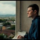 Ein Mann steht seitlich an einem offenen Fenster, trägt eine blaue Jacke und blickt auf eine Stadtlandschaft.