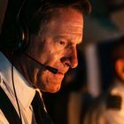 Ein fokussierter Pilot im Cockpit mit Headset, in warmer Beleuchtung sitzend. Ein weiterer Pilot ist im Hintergrund zu sehen.