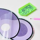 Lila kreisförmige Vorlage mit schwarzen Punkten, grünes Tablett voller kleiner, blauer Perlen, daneben ein rosa Stift.