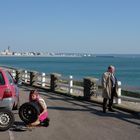 Nummernschild: 1434 RD 76. Eine Frau wechselt einen Reifen an einem roten Auto. Ein Mann steht am Meeresspaziergang.
