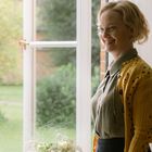 Eine lächelnde Person steht neben einem offenen Fenster mit hellem, weichem Vorhang. Draußen ist Grün zu sehen.