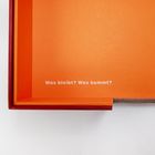 „Was bleibt? Was kommt?“ auf orangefarbenem Hintergrund. Eine geöffnete Schachtel mit roten Kanten.
