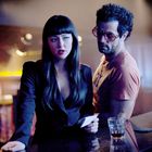 Zwei Personen in einer Bar. Eine Frau im schwarzen Blazer hält Papier, ein Mann in orange trägt Sonnenbrille.