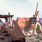 Drei Personen sitzen in einer Planierraupe, eine Person steht mit einem Gewehr auf der Schaufel. Wüstenlandschaft im Hintergrund.