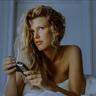 Kein Text erkennbar. Eine Frau mit langen, blonden Locken hält eine Fernbedienung, im Hintergrund eine Wandlampe.