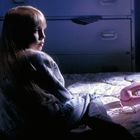 Ein Kind sitzt auf einem Bett und blickt nach links. Auf dem Bett liegt ein rosa Telefon. Die Szene ist beleuchtet.