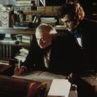 Zwei Männer in historischer Kleidung in einem Büro; einer schreibt bei Kerzenlicht, während der andere über seine Schulter schaut.