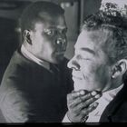 MNL-73B. Ein Mann in Anzug hält sanft den Kopf eines anderen Mannes und schaut ihm ins Gesicht.